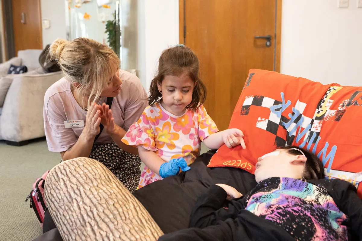 A young girl and a Martin House care team member sit with a child resting on a sofa, playing together during a session at the hospice