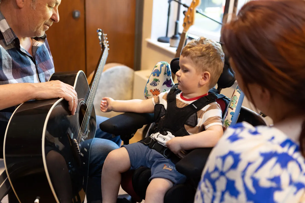A music therapist plays acoustic guitar for a young boy in a supportive wheelchair at Martin House Children’s Hospice, as a carer sits alongside them