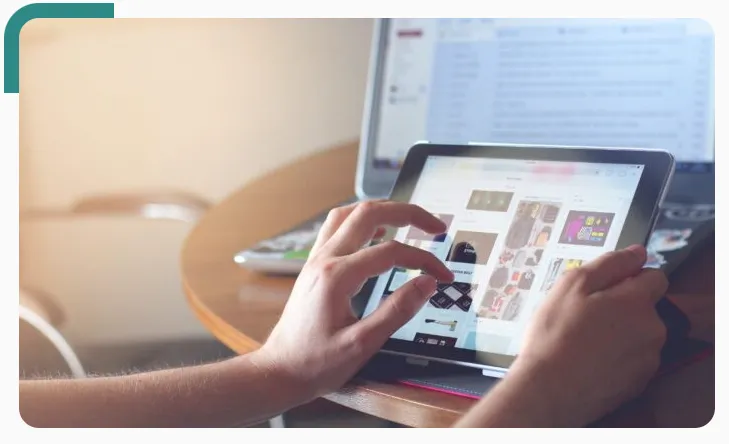 Hands browsing a product catalogue on a tablet, a laptop open behind it